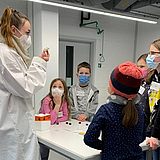 Eine Person mit Maske erklärt den Kindern ein Experiment