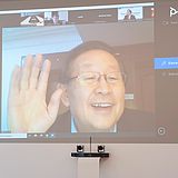 Eine Person sitzt vor einer großen Projektionsfläche, die eine Online-Videokonferenz zeigt