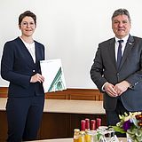 Eine Person hält ein Dokument in der Hand und steht neben einer Person