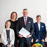 Person hält eine Urkunde in der Hand und steht neben ihrer Familie und anderen Personen